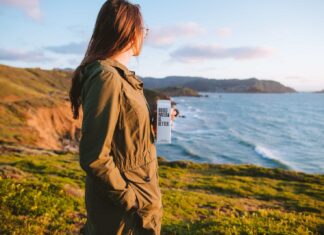 woman in brown jacket standing on green grass field near body of water during daytime