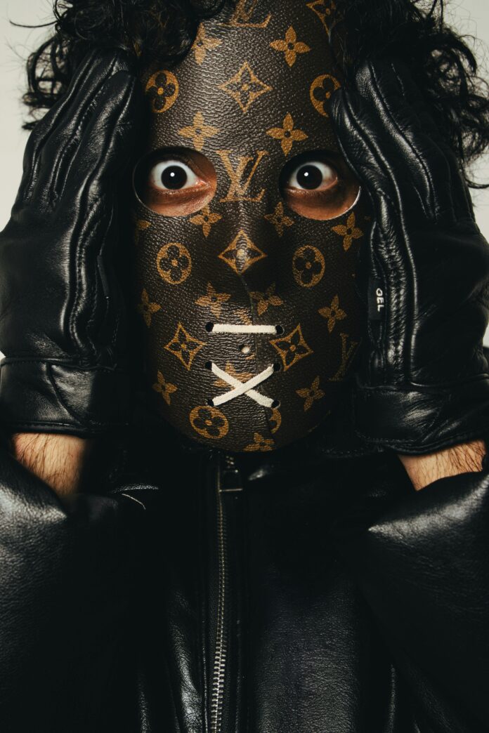 Close-up of a person in a mysterious monogram Louis Vuitton leather mask and black gloves, holding their head with a wide-eyed, shocked expression.