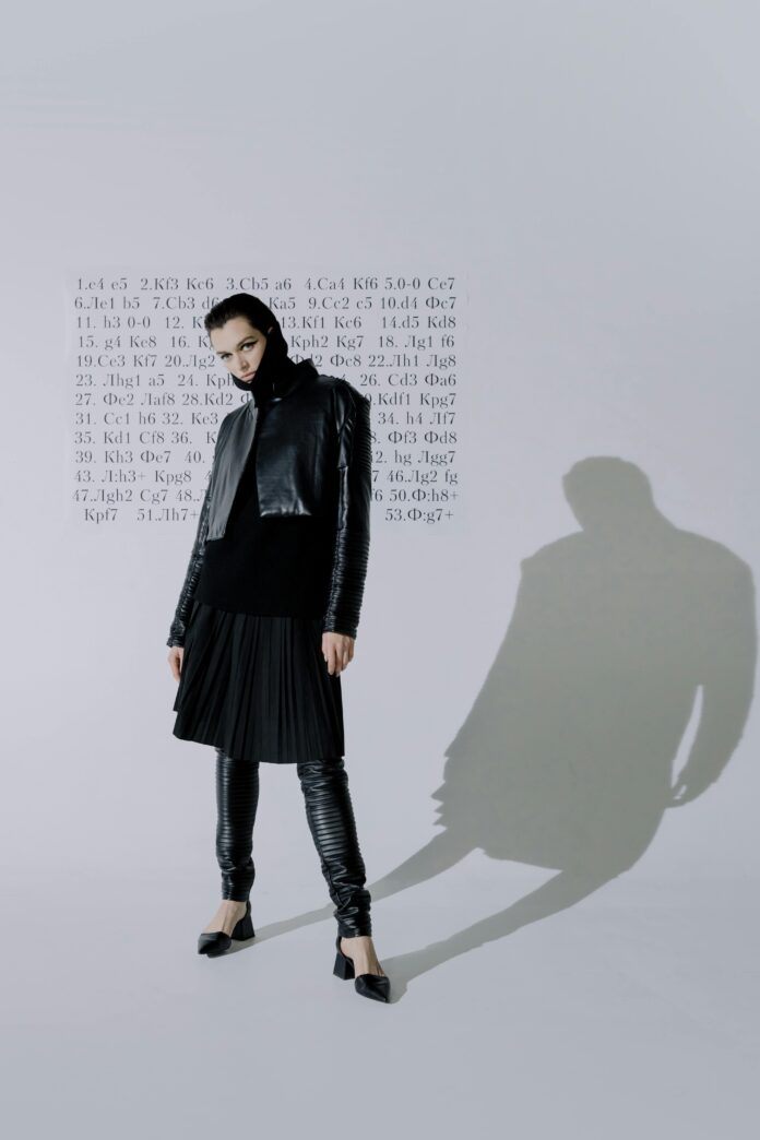 Model dressed in all -black posing against a white background with a long text of letters and symbols behind her